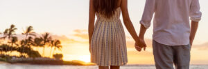 Romantic couple holding hands watching sunset on tropical beach with palm trees and ocean waves.